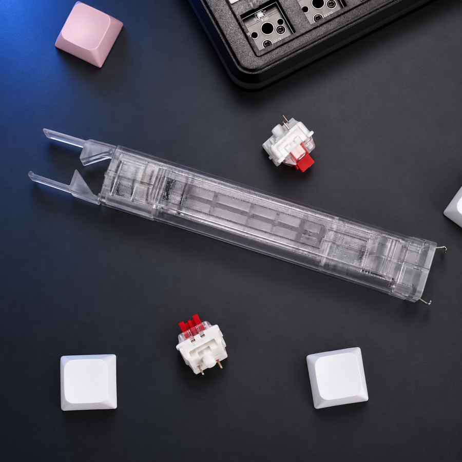 Clear Lift transparent two-in-one keycap and switch puller on a charcoal studio surface. Surrounding keyboard, switches and loose keycaps are not included.