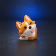 Corgi Charm orange and white resin keycap with pink tongue, angled view on a charcoal studio surface with blue lighting.